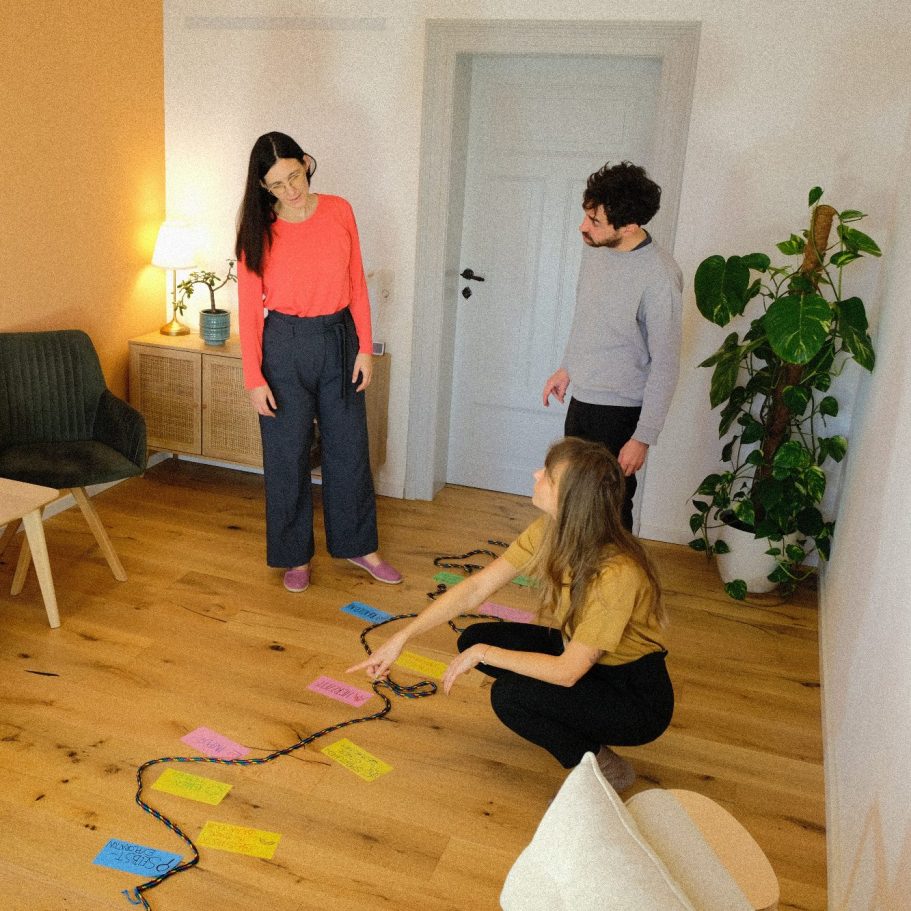 Drei Personen - die Beraterin und ein Paar - arbeiten in einem Raum. Auf dem Boden liegt ein Seil mit Moderationskarten.